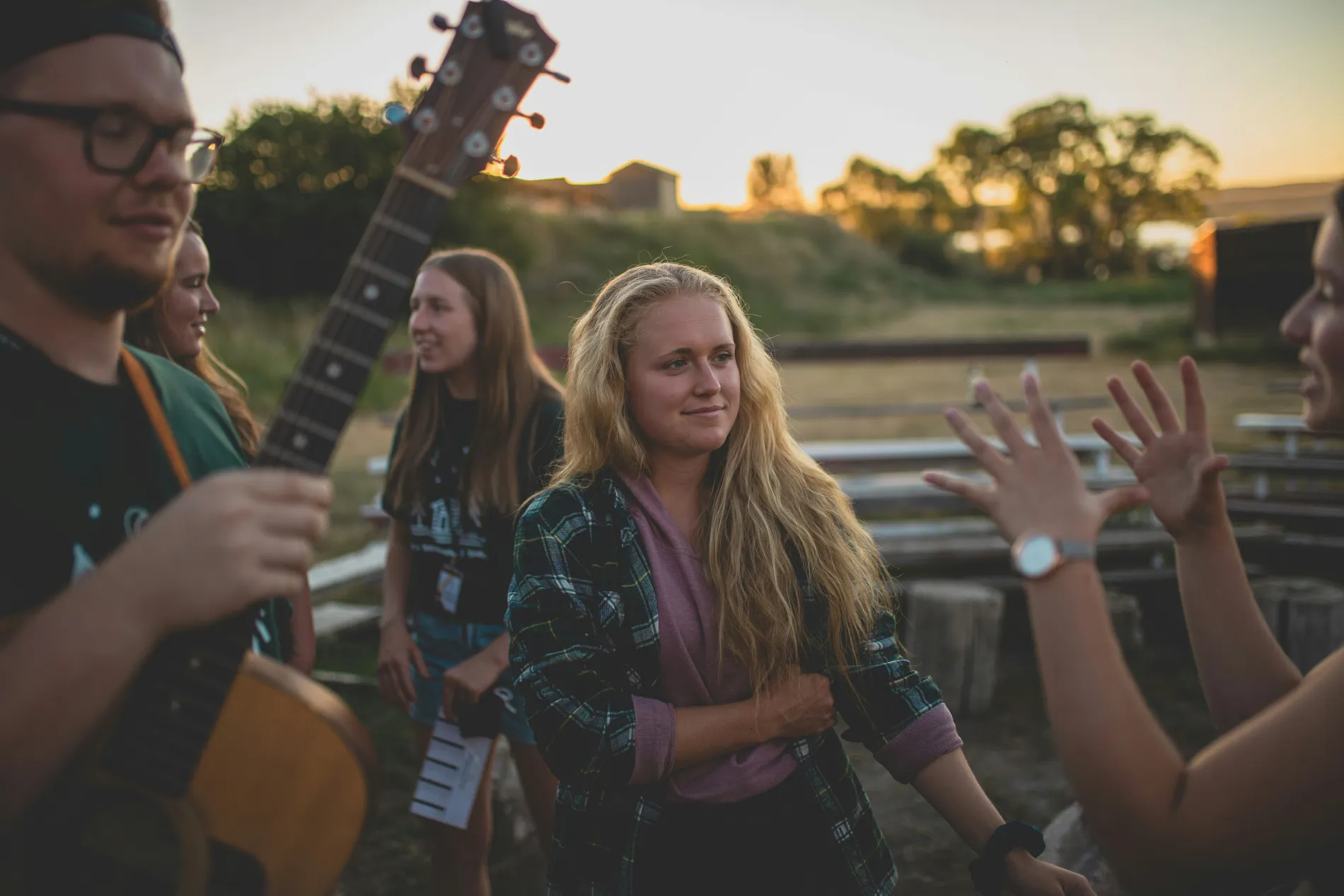 A group of young musicians laughing together outdoors at sunset, one holding an acoustic guitar